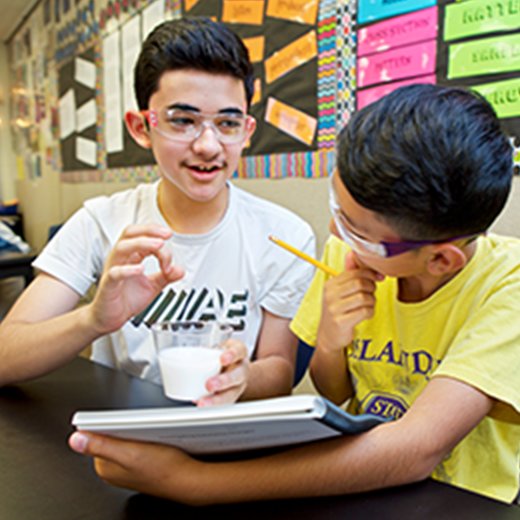 Two middle school students, one holding a beaker and a tablet, engaged in a science experiment in an 