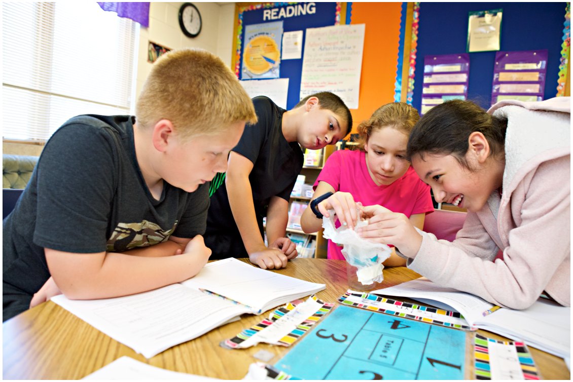 Four middle school science students gather around a desk, engaged in an interactive group project, investigating an object together in a colorful classroom setting.