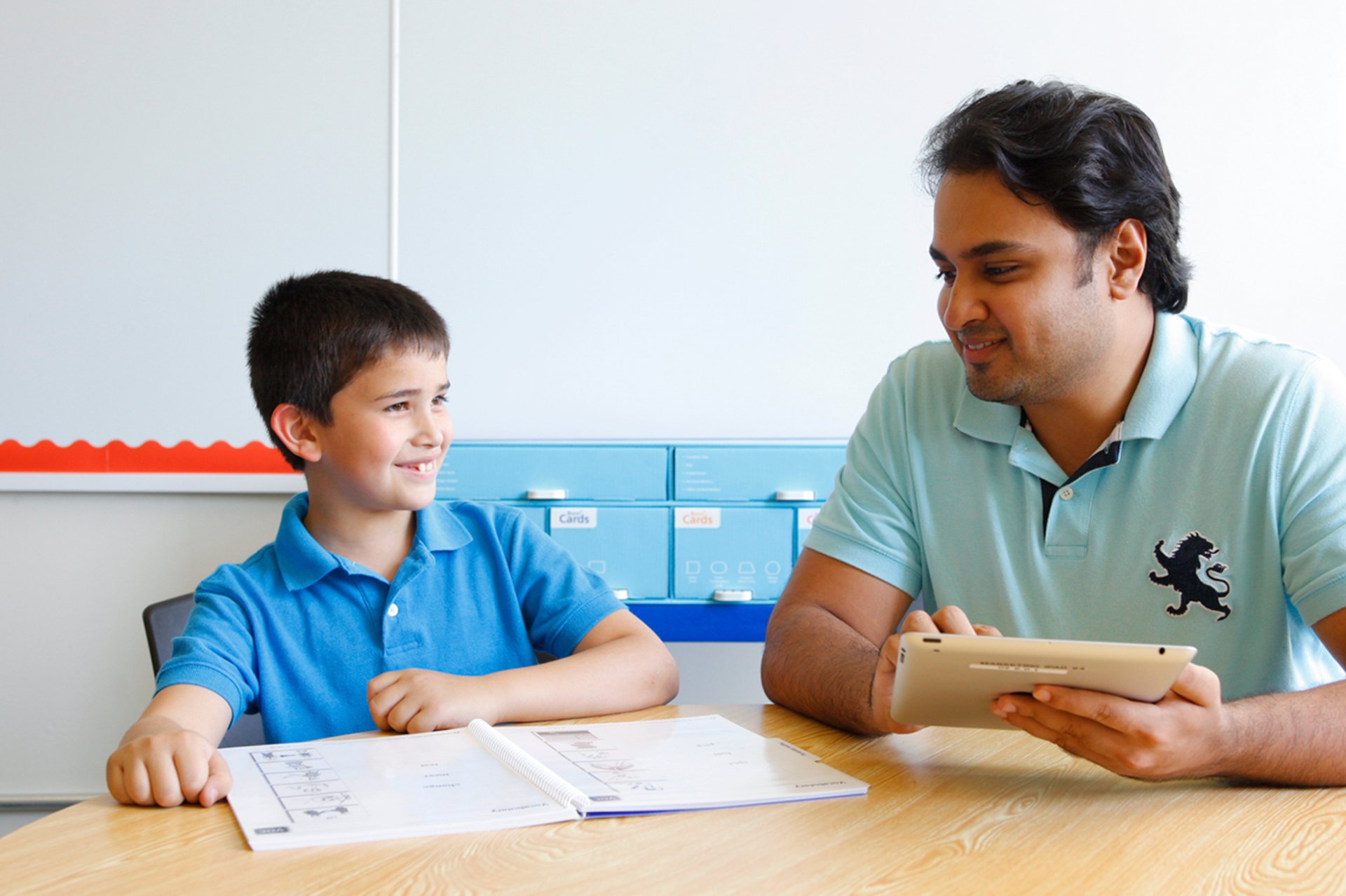 A man and a young boy in blue polo shirts sit at a table in a classroom, interacting and smiling as the man programs a tablet.