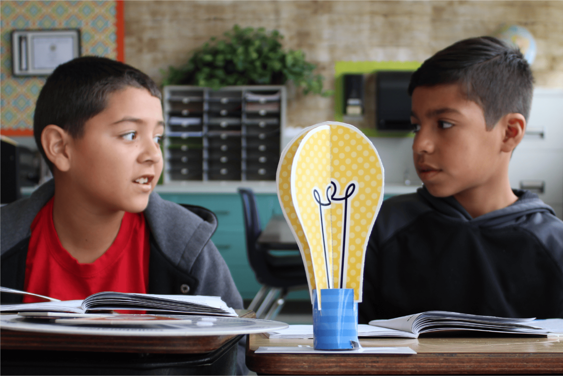 Two boys in a classroom setting, one looking at the other who is speaking, with a lightbulb decoration and school supplies on their desk as they engage in an online language arts curriculum.