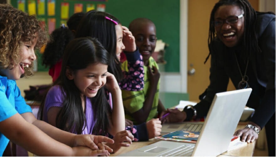 A diverse group of young students and a teacher laughing together around a laptop in a colorful classroom, engaged with an online language arts curriculum.
