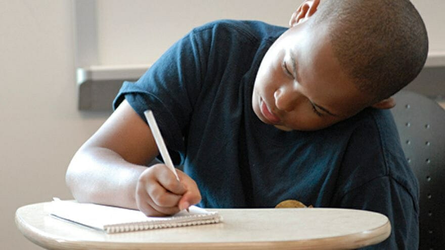 A young boy concentrating on his online language arts curriculum while writing in a notebook at a desk.