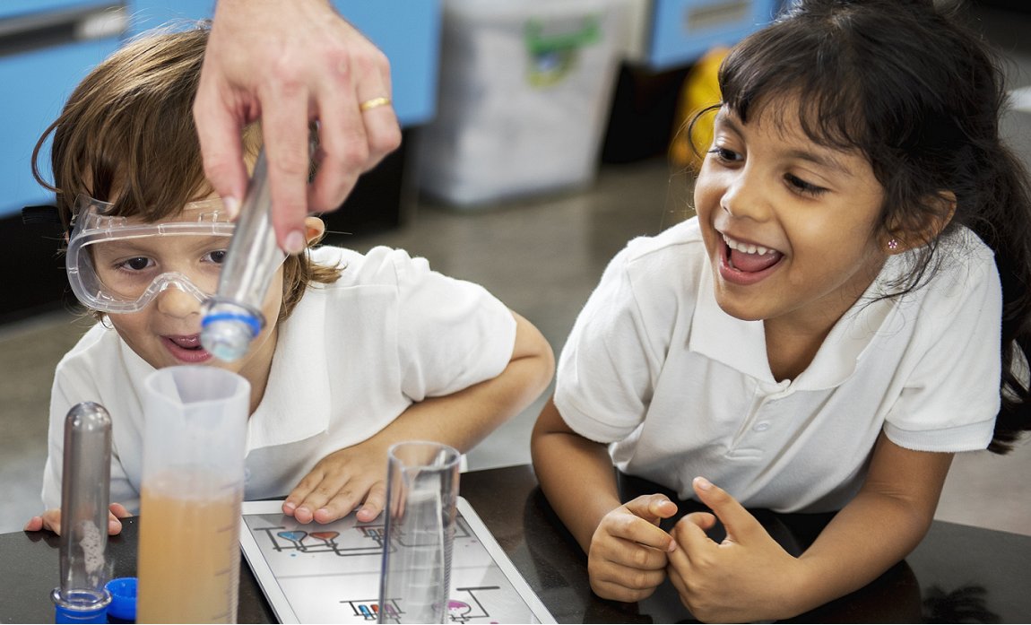 Young girl in goggles watches with excitement as an adult's hand demonstrates a science experiment with test tubes.