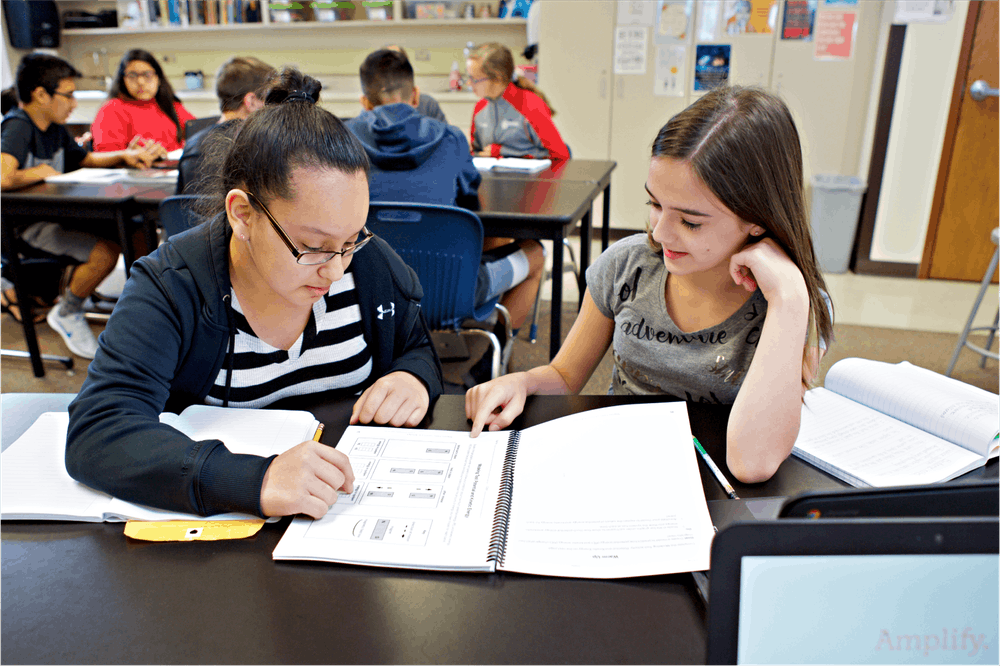 Two students, one using glasses, collaboratively studying from a textbook at a classroom desk with other students in the background.