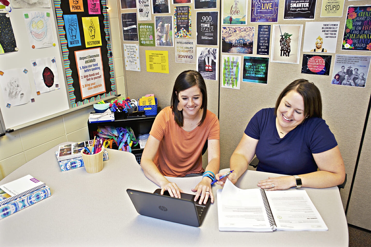 Two women smiling and working together at a desk with a laptop and notebook, surrounded by colorful posters on the walls.