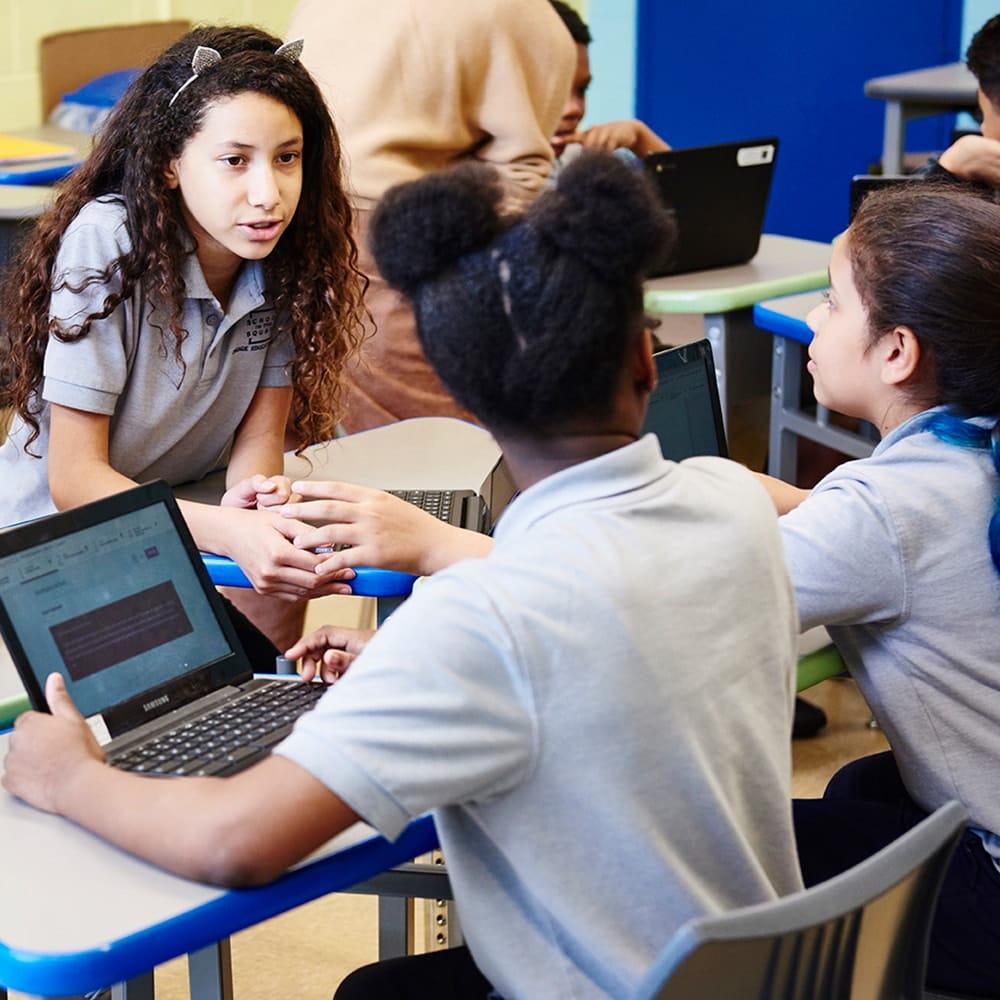 Two children sit at a table with Chromebooks, facing each other and talking, in a classroom with books and storage bins in the background.