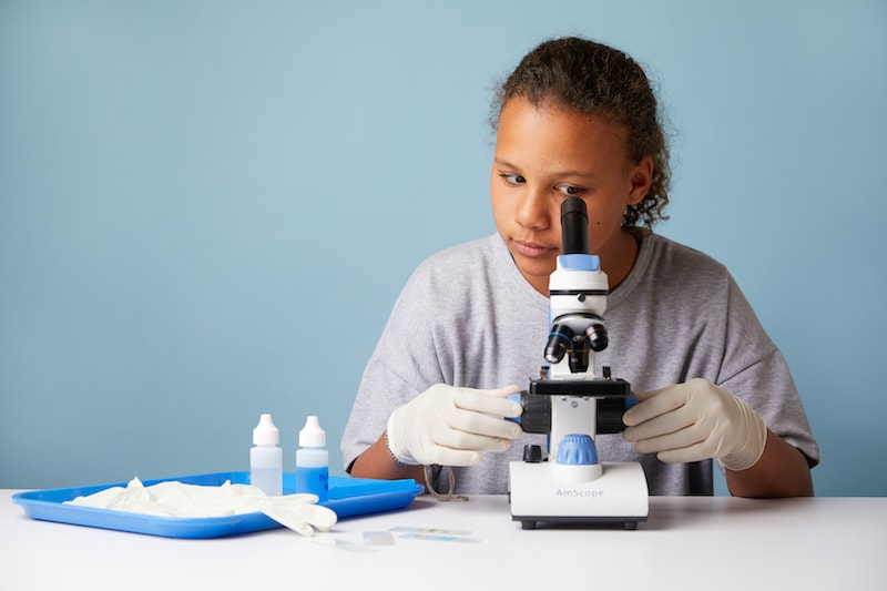 A young girl wearing gloves uses a microscope, with scientific tools and samples on the table in front of her, against a light blue background.