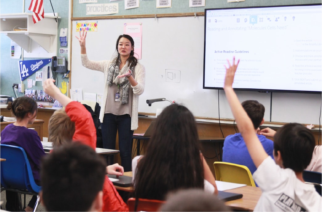 A teacher discusses educational content in front of a whiteboard while students in the classroom raise their hands eagerly.