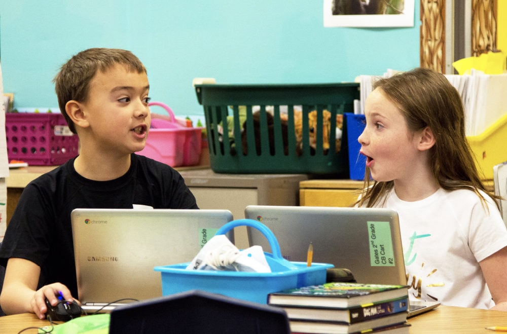 Two children engaged in an animated conversation while sitting at a school desk with laptops and books.