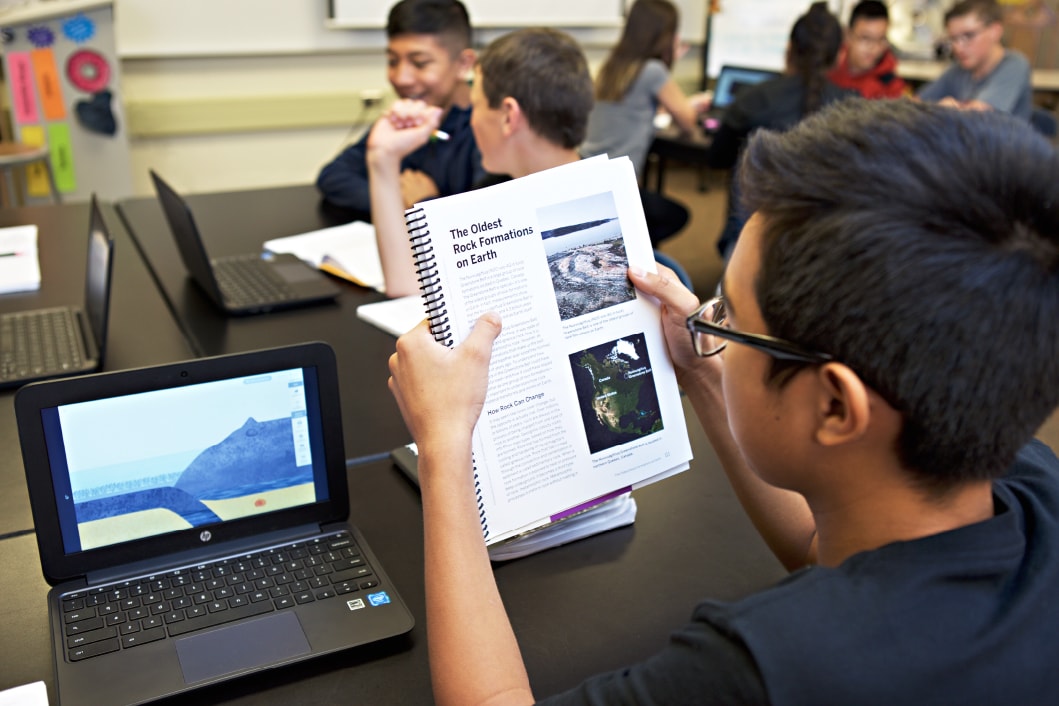 A student in a classroom holding up a textbook about fossils, with a laptop open in front of him and classmates in the background.