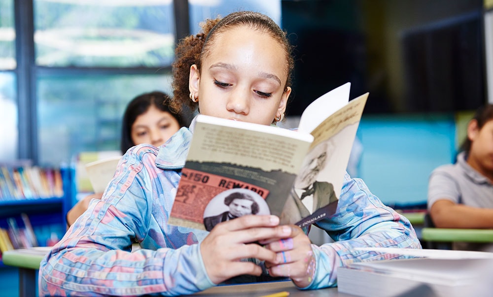 A young girl reading a book intently in a classroom with other students blurred in the background.
