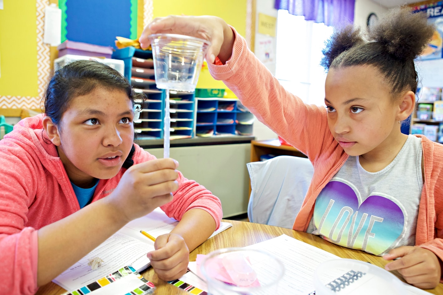 Two students conducting a science experiment in a classroom, one pouring liquid into a container while the other observes.