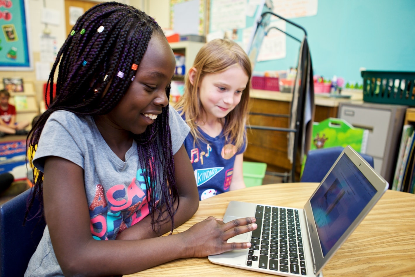 Two young girls, one black and one caucasian, smiling and using a laptop together in a colorful classroom setting.