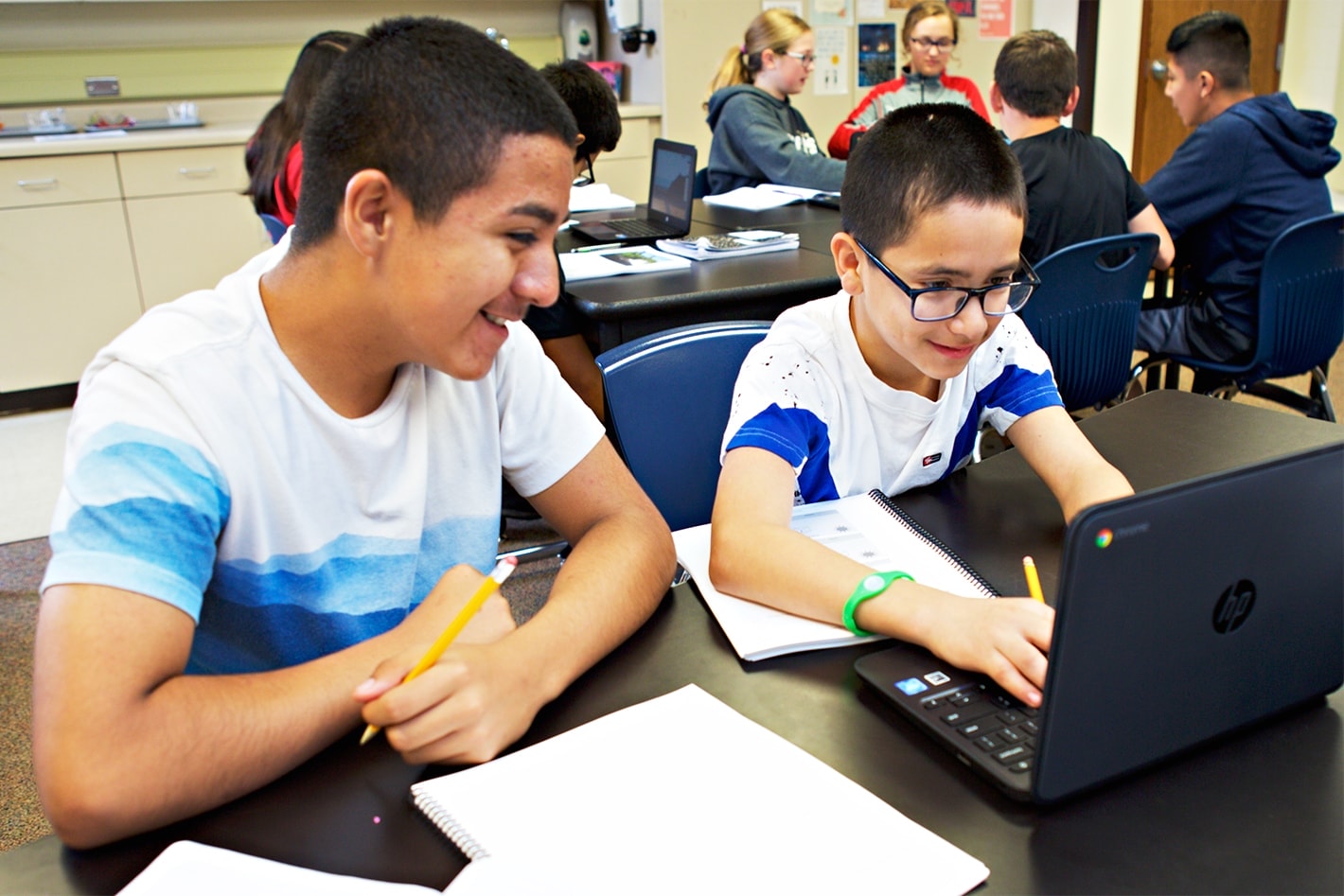 Two students, one using a laptop and the other writing in a notebook, smiling and working together in a classroom.
