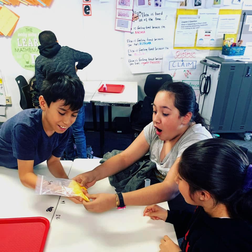 Three students around a table engaged in a group activity with papers and markers, in a classroom setting with educational posters on the wall.