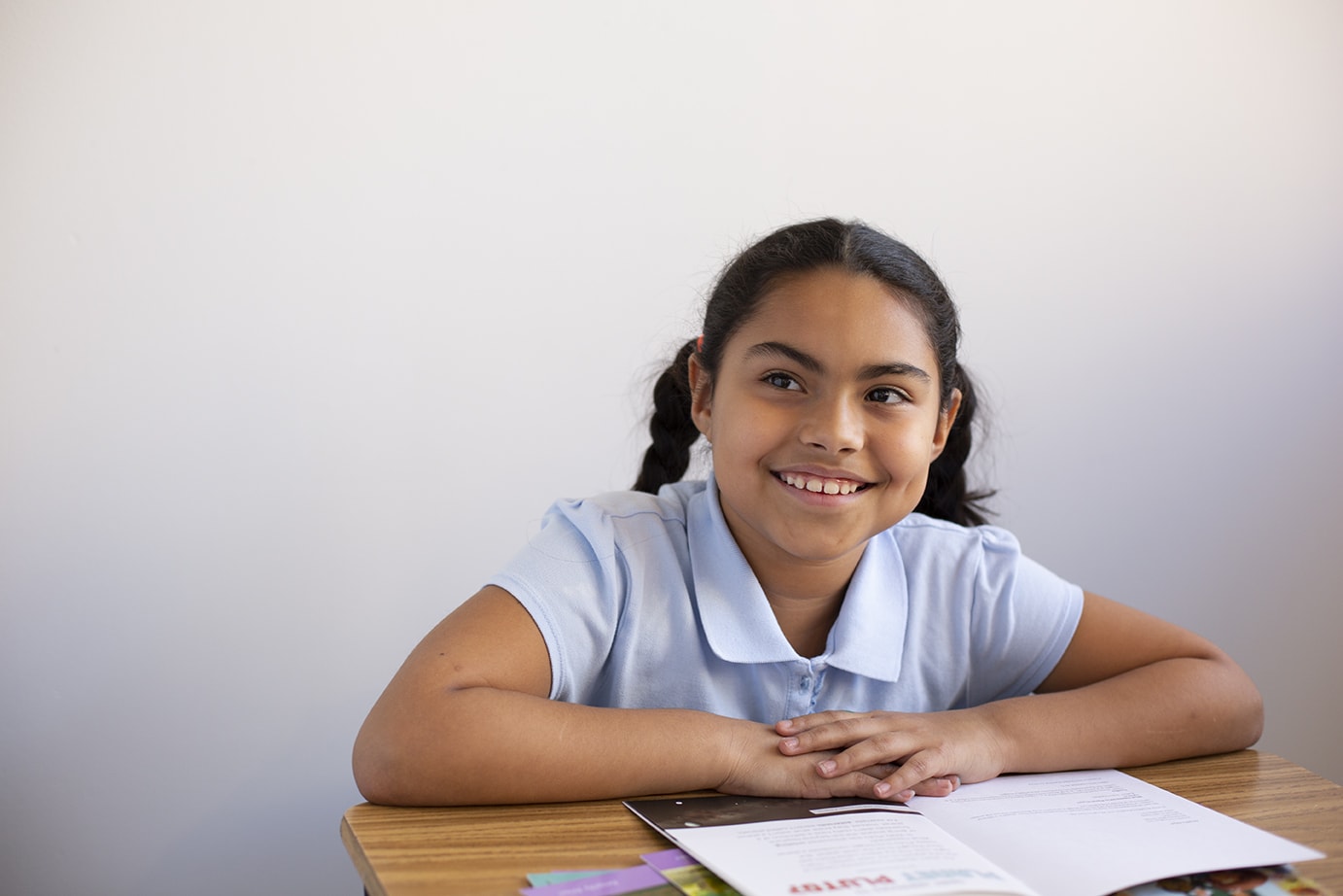 Young girl smiling at camera while sitting at a desk with books, wearing a light blue polo shirt.