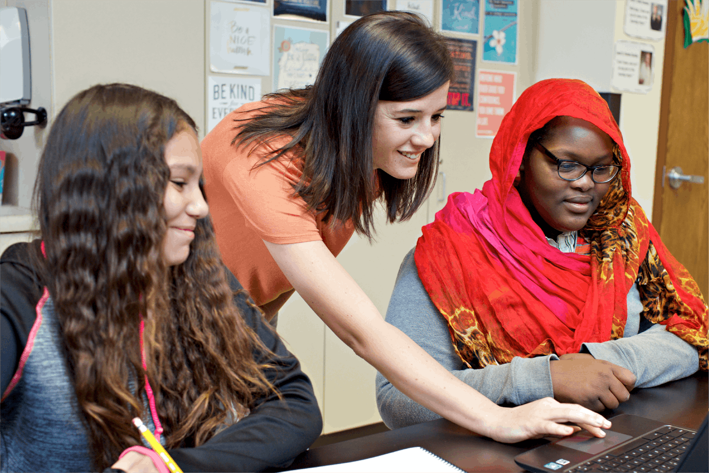 Teacher assisting two students, one wearing a hijab, with a laptop in a colorful classroom setting.