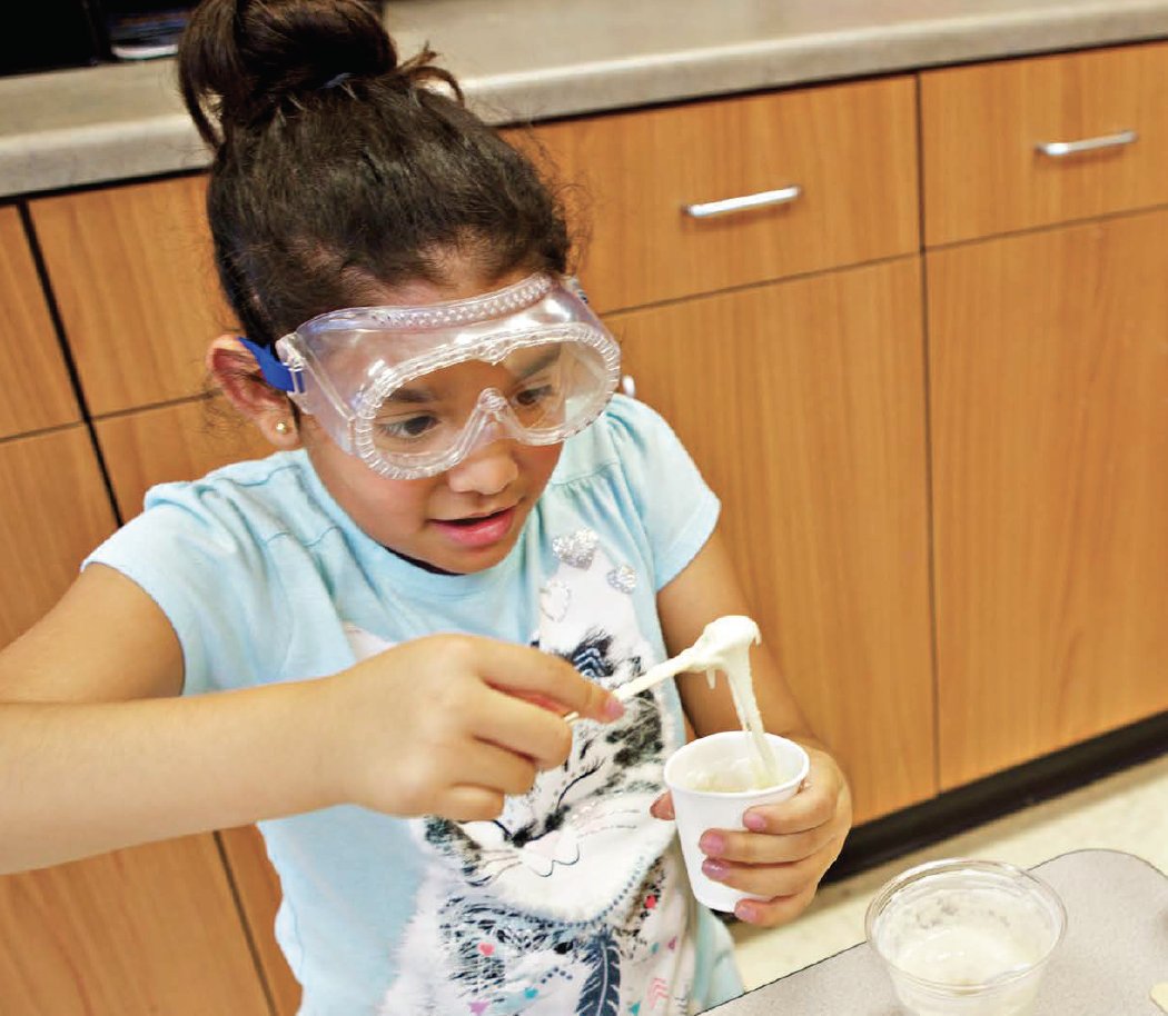 A young girl wearing safety goggles conducts a science experiment, stirring a cup with a pipette in a classroom setting.