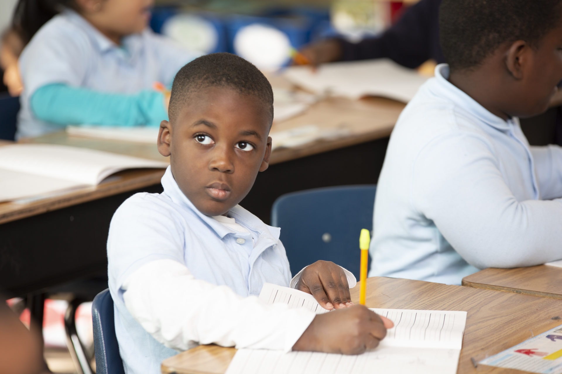 Young boy in a classroom, holding a pencil, looking at the camera while classmates work in the background.