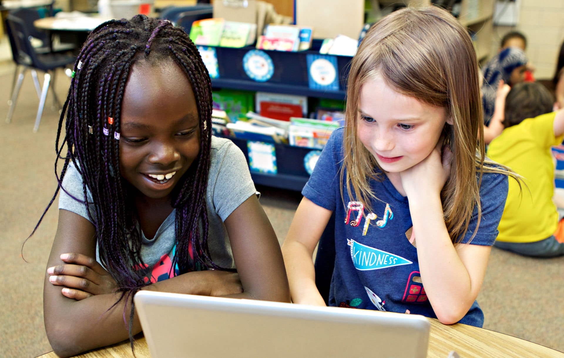 Two young girls, one with braided hair and the other with blonde hair, smiling and looking at a laptop screen in a classroom.