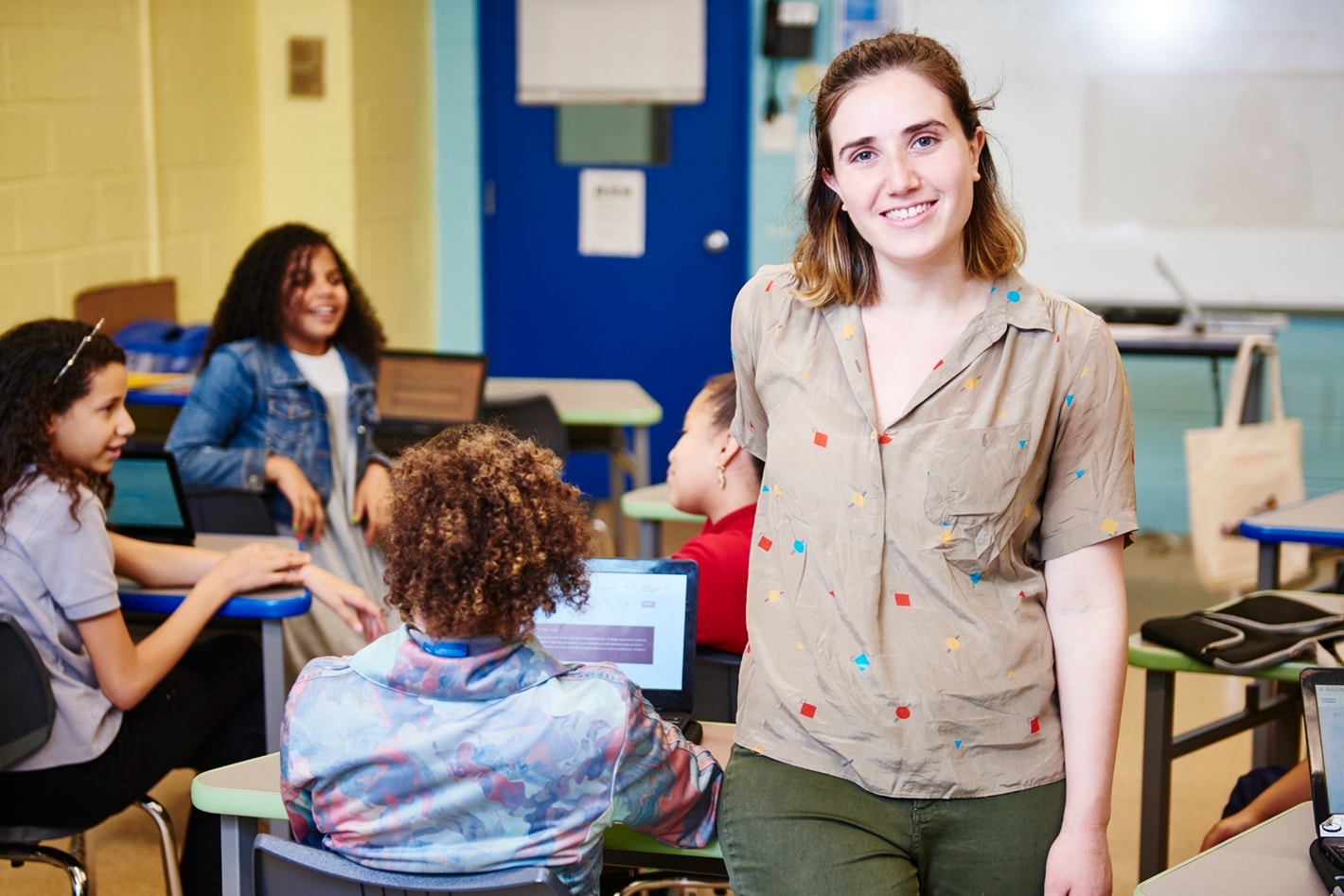 A female teacher smiling at the camera in a classroom with diverse students engaged in background activities.