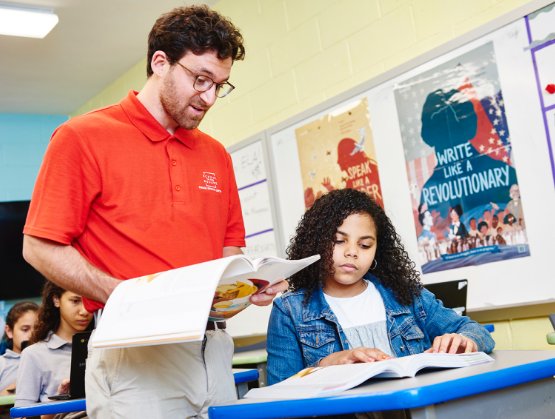 A male teacher in a red shirt explains a textbook to a young female student at her desk in a colorful classroom.