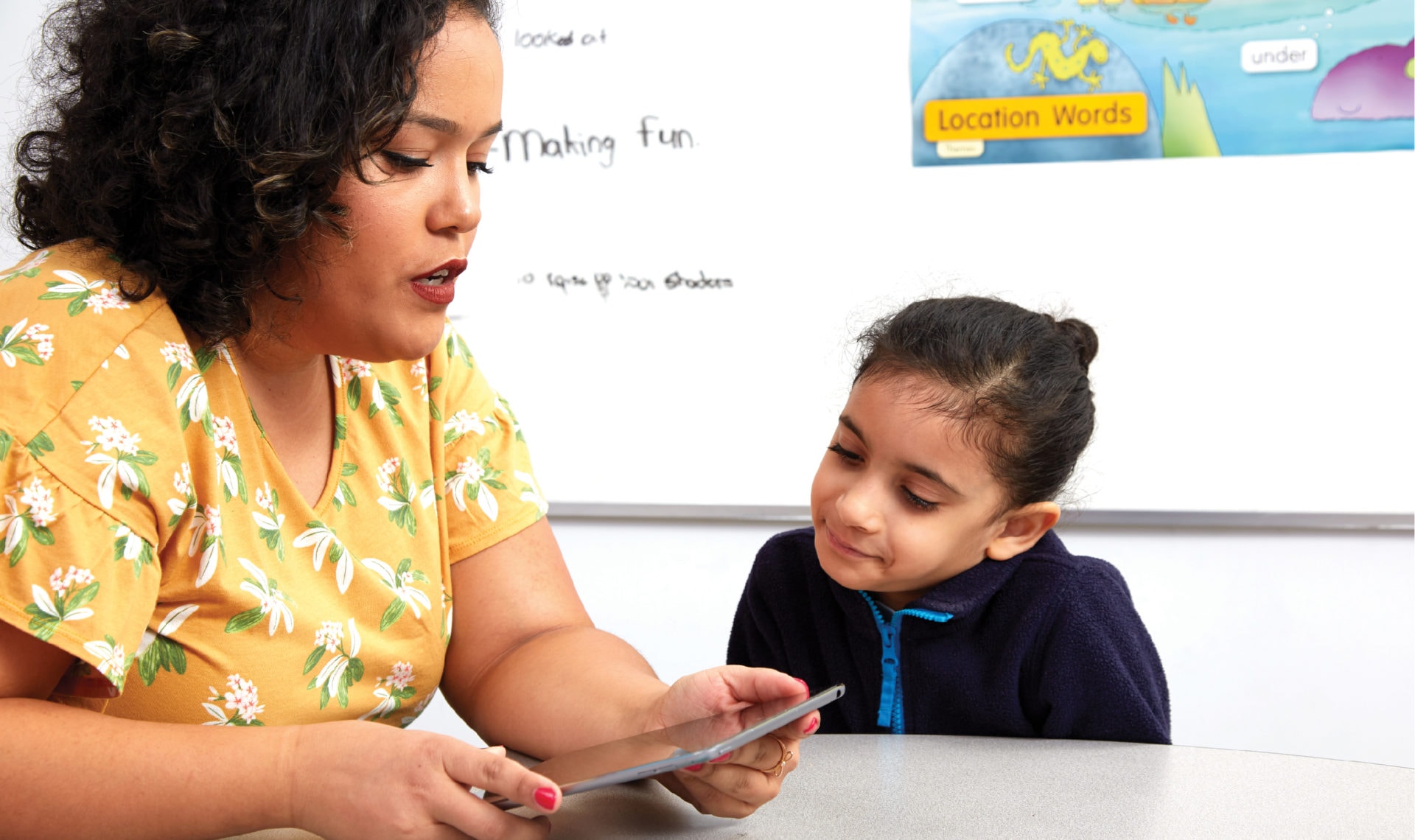 A teacher using a tablet interacts with a young student in a classroom, with educational posters in the background.