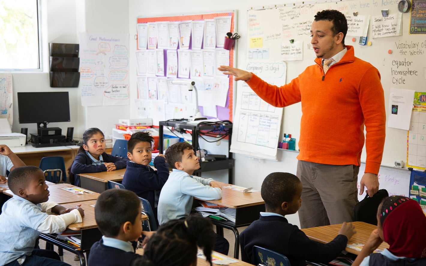 A teacher in an orange sweater gesturing while explaining a lesson to attentive elementary students in a classroom.