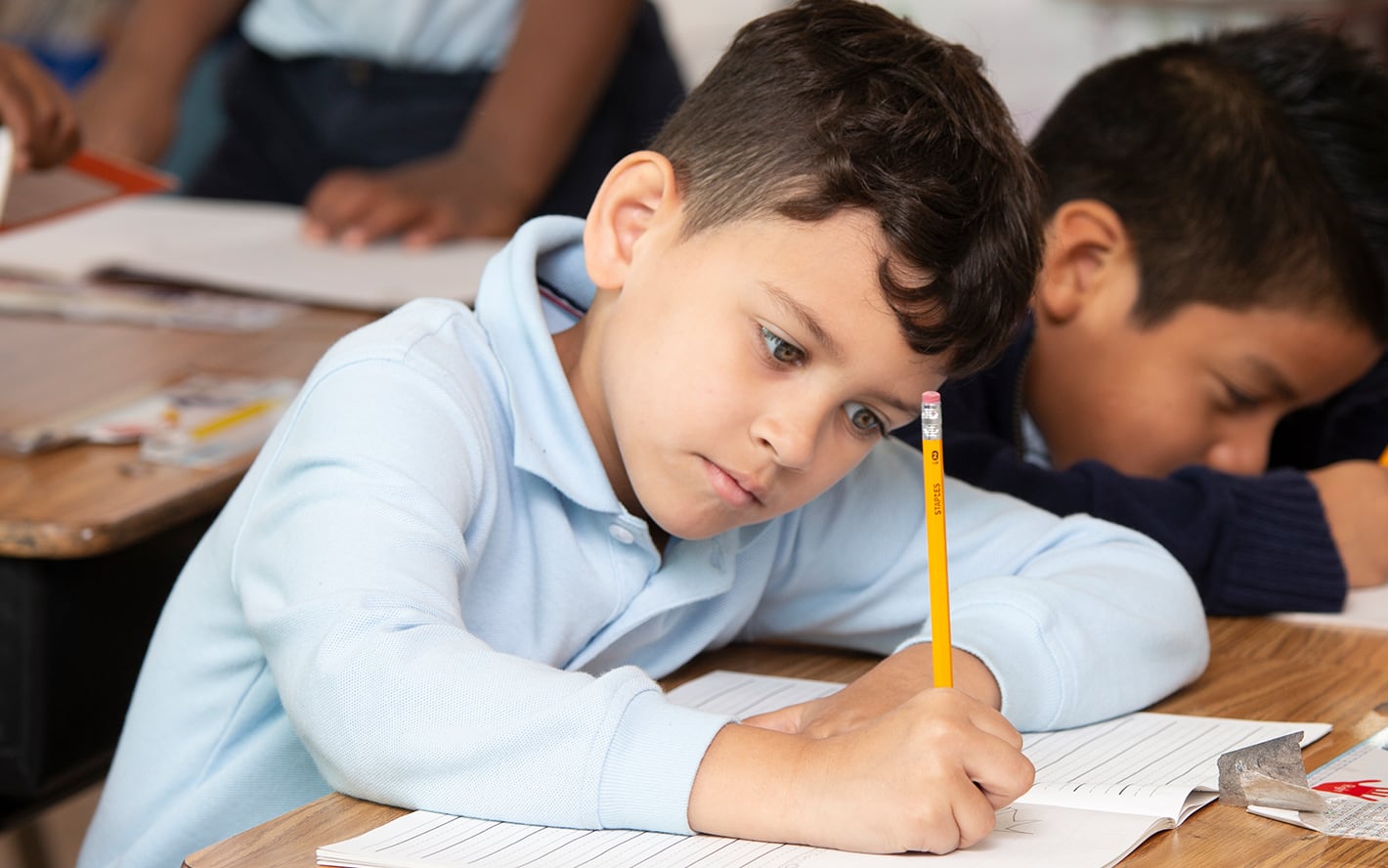 A young boy in a blue shirt focused on writing in a classroom, holding a pencil, with another student in the background.