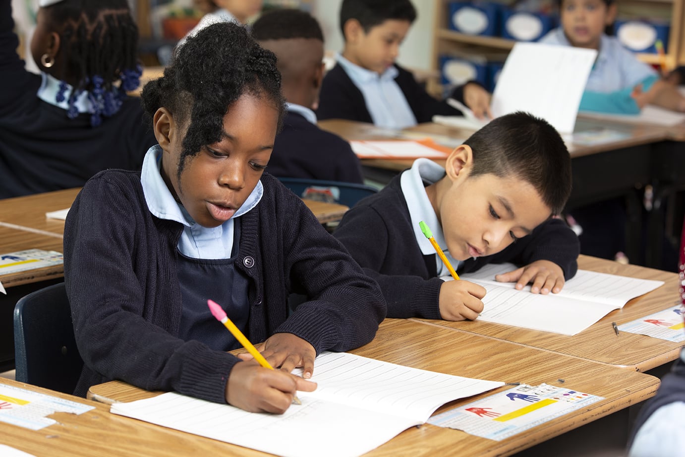 Two students in school uniforms focused on writing in their notebooks at a classroom desk during a science lesson.