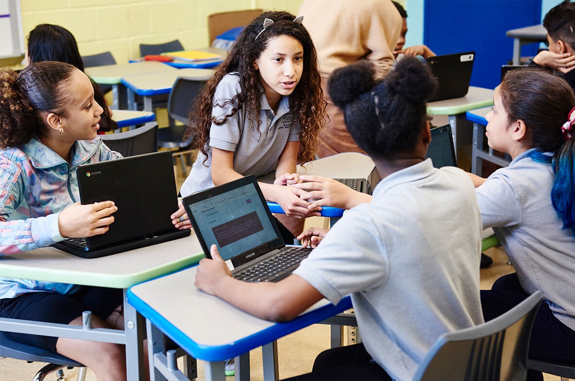 Group of diverse middle school students working on laptops and discussing in classroom.