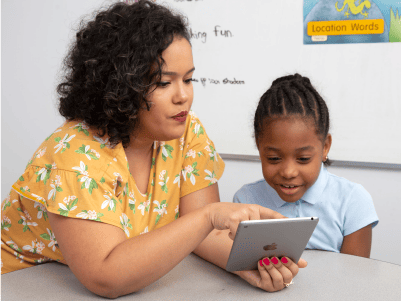 A woman and a young girl look at a tablet together in a classroom, with educational posters visible in the background.