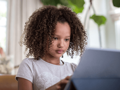 Young girl with curly hair using a tablet in a bright, plant-filled room.
