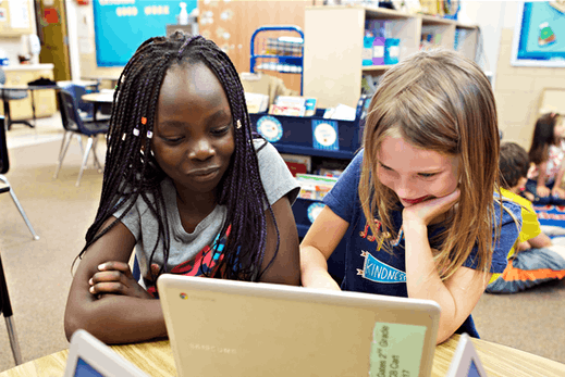 Two young girls, one black and one Caucasian, smiling and looking at a tablet together in a colorful classroom, engaged in an online reading program.