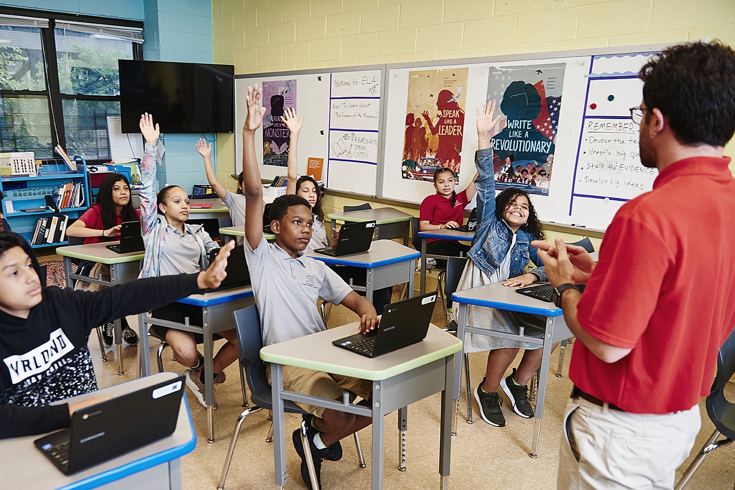 A diverse group of students raising their hands in a classroom, engaging with a male teacher who is standing in front of them.