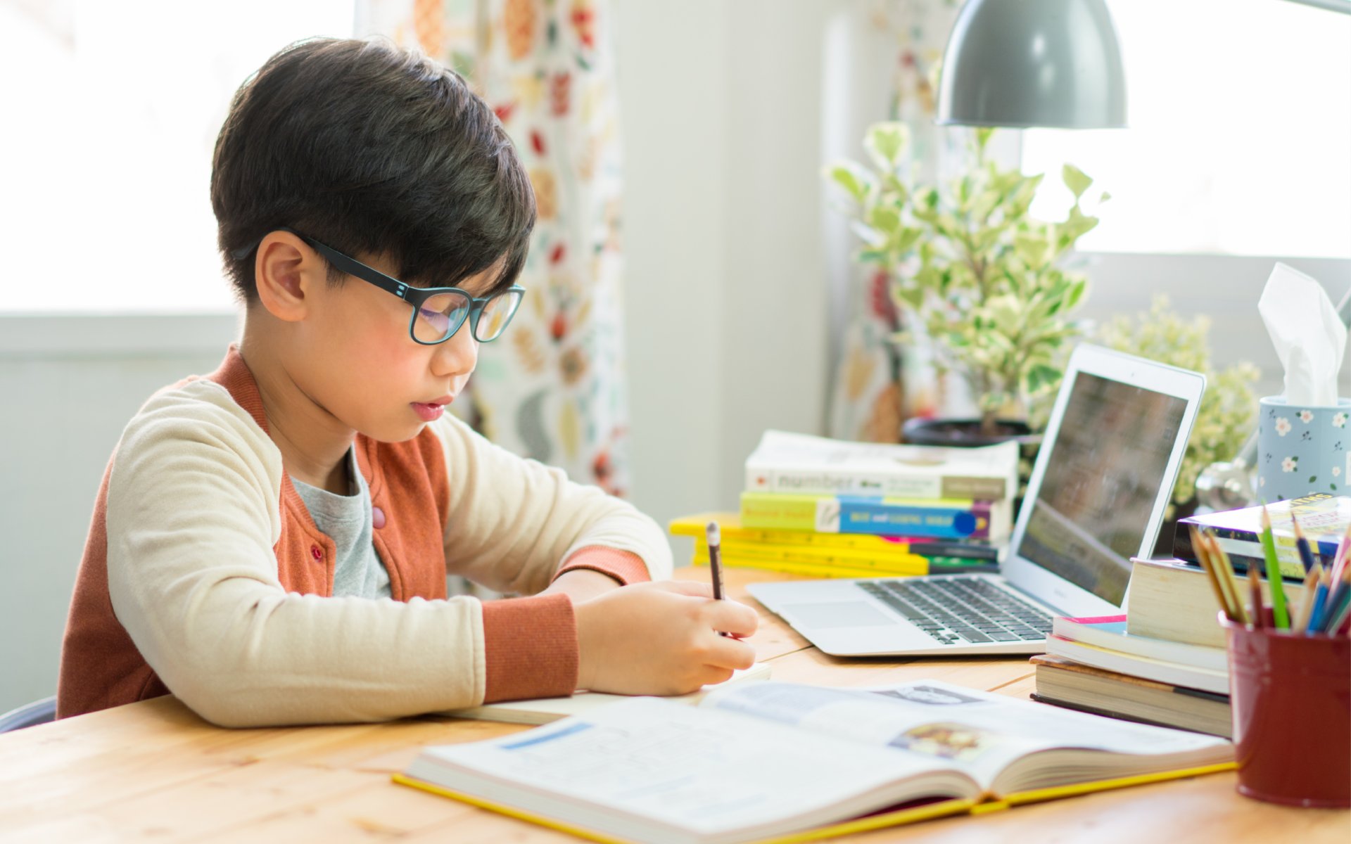 Young boy in front of a textbook and laptop in a living space writing something