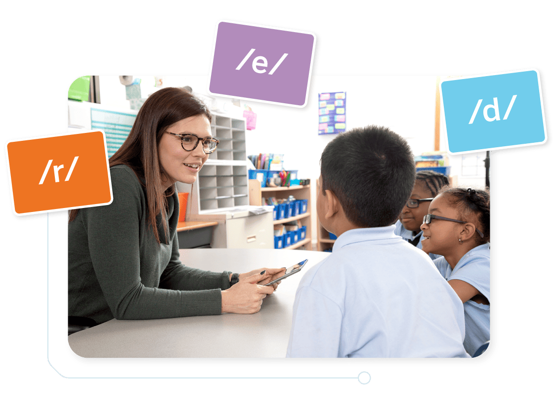 A teacher interacts with three young students at a desk, using flashcards to teach phonetics in a classroom.