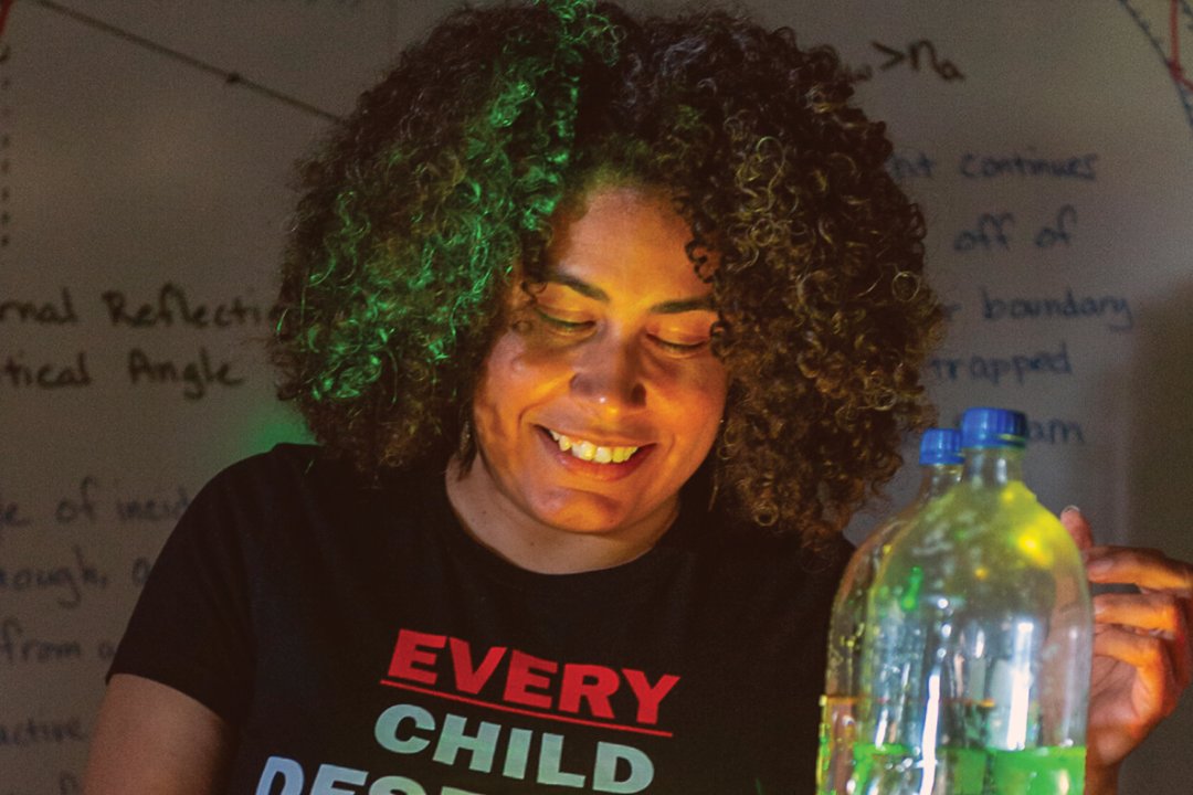 A woman with curly hair, smiling at a glowing bottle in a dark classroom, with scientific equations written on a blackboard behind her, relates to Juan Vivas, a noted SpaceX engineer.