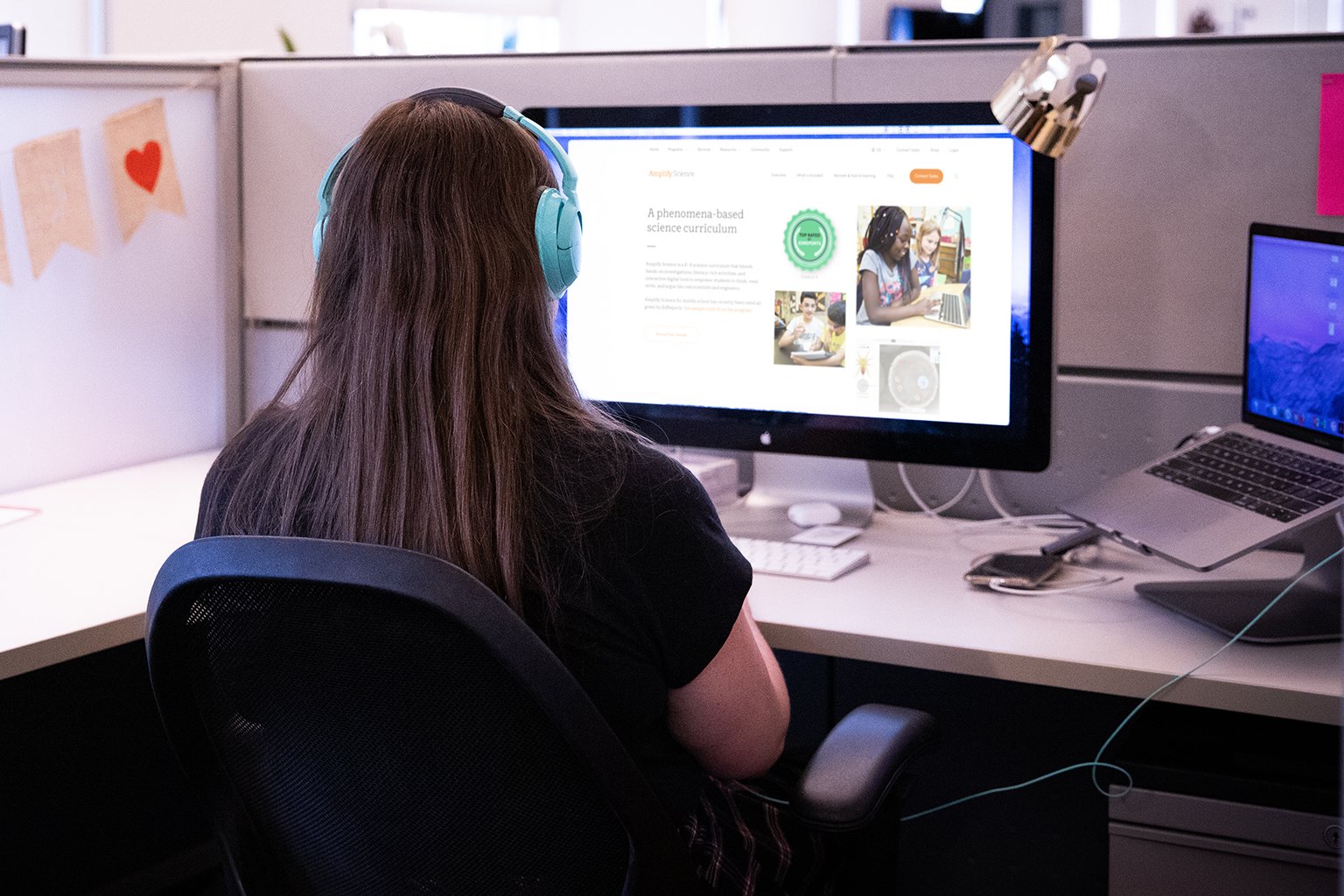 A woman wearing headphones works at a desk with two monitors displaying content-related websites.