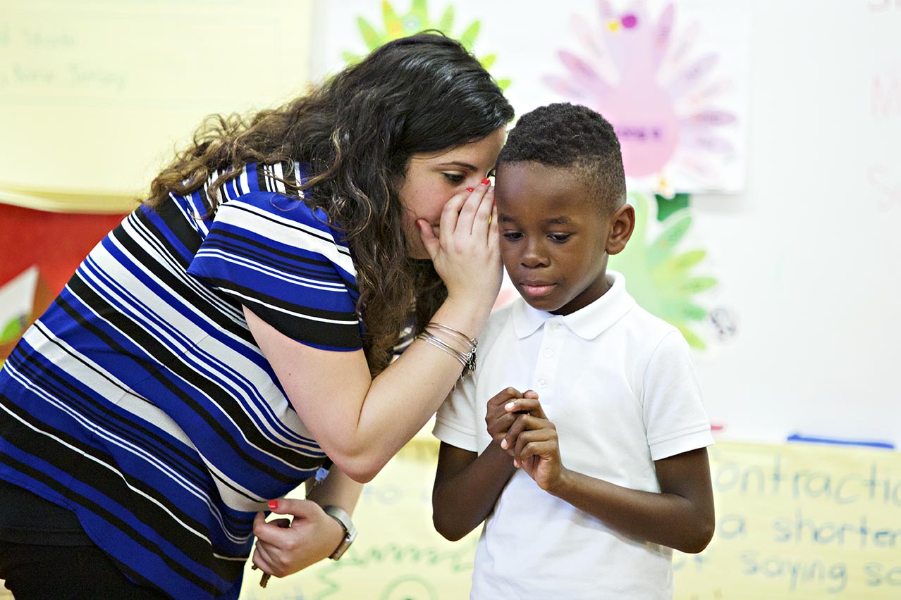 A woman whispering to a young boy in a classroom, with educational posters visible in the background.