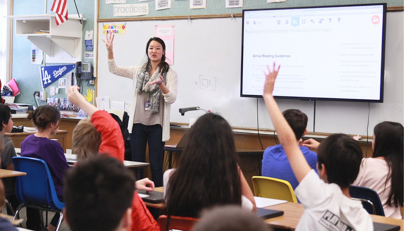 A math teacher stands in front of a classroom, engaging with students who are raising their hands, with a digital board displaying 