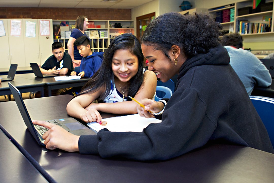 Two students, one using a laptop and the other taking notes, are smiling and engaging together at a table in a classroom.