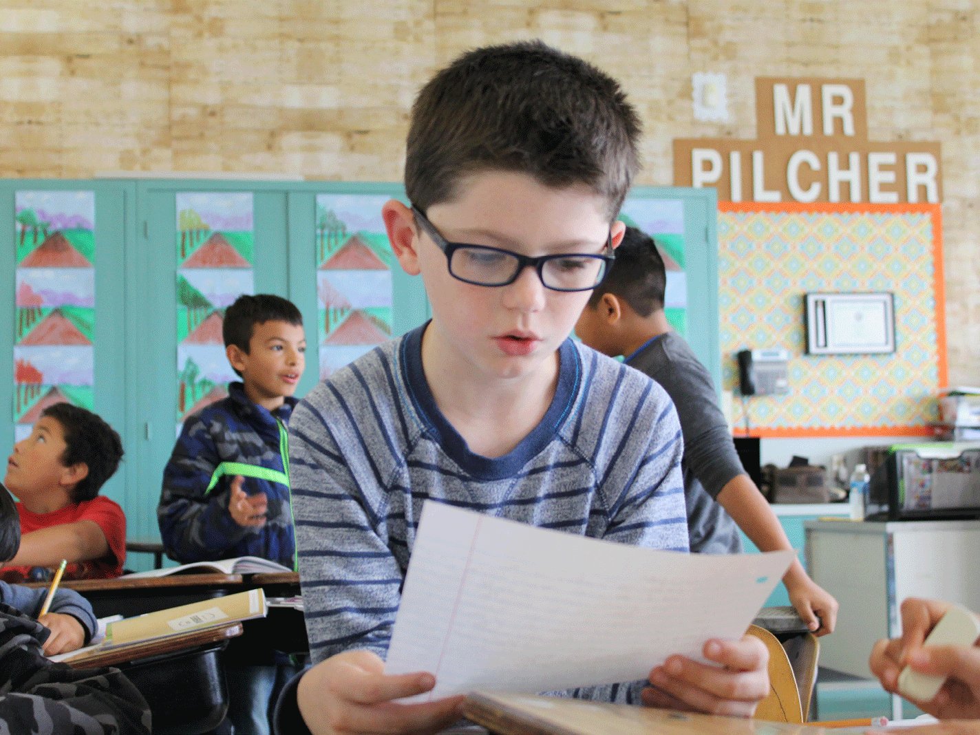 A boy in glasses improves his reading with a paper in a classroom among other students and colorful art in the background.