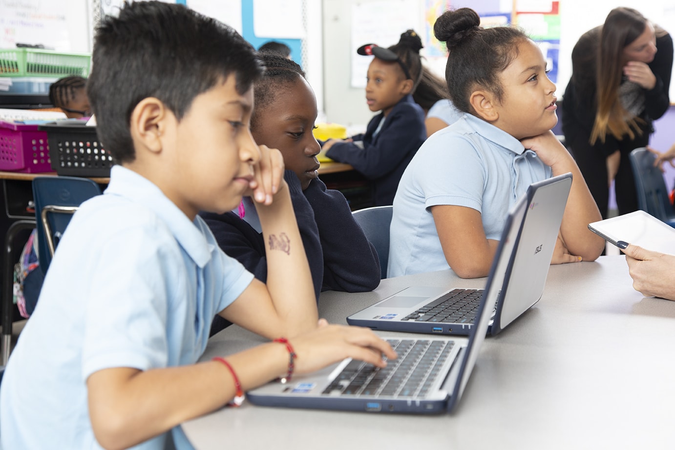 Elementary school students using laptops in a classroom, intently focused on their screens while a teacher assists others in the background.