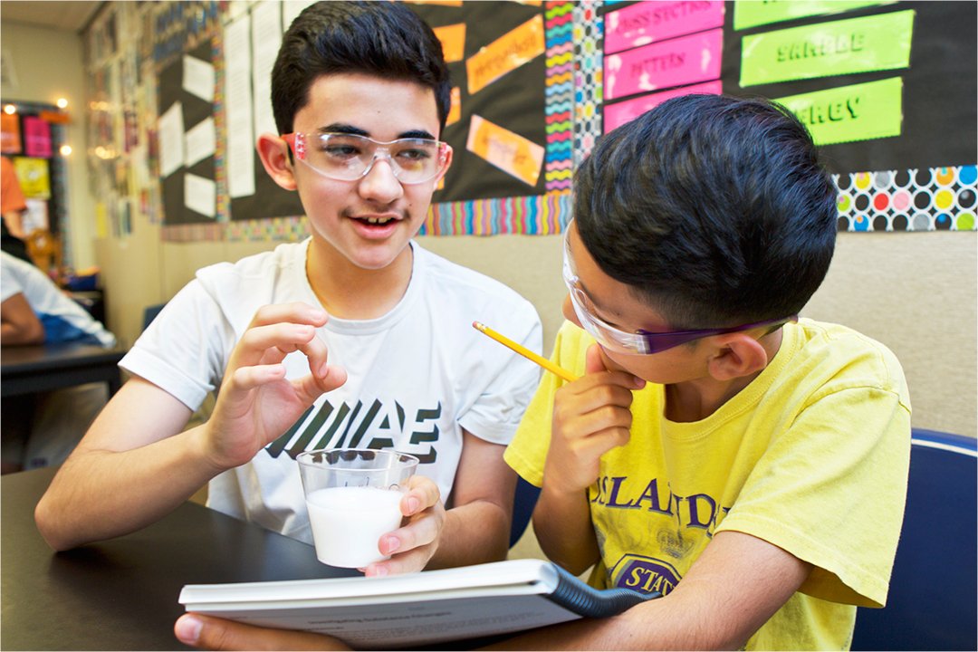 Two teenage boys engaged in a science experiment in a classroom, one holding a magnifying glass and the other explaining how to engage students.