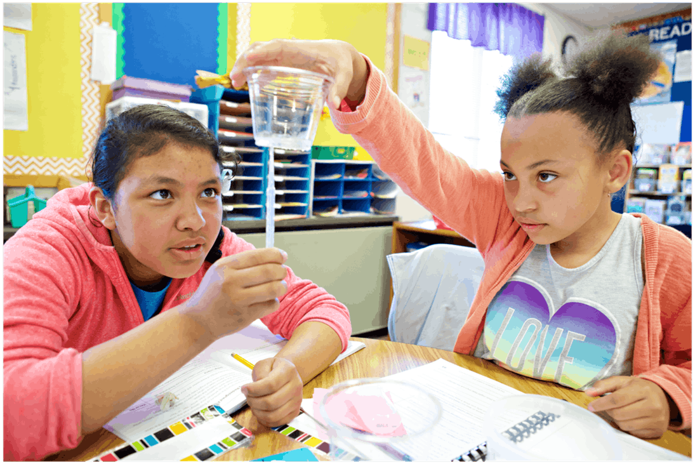 Two students engage in a science experiment in a classroom, with one holding a cup of water and a stick while the other observes intently.