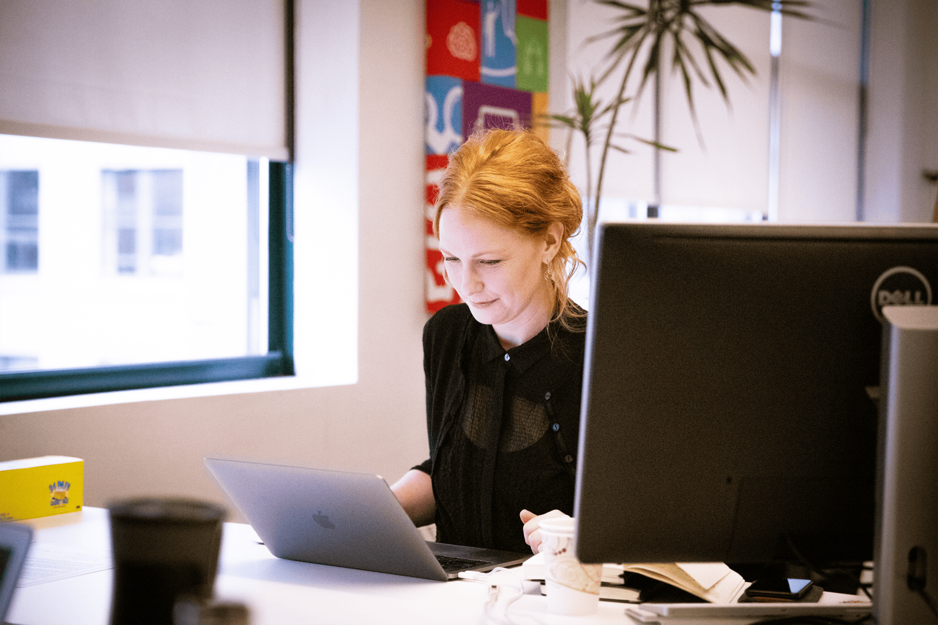 A woman with red hair working on a laptop at an office desk, with a desktop monitor beside her and posters advertising Amplify Tutoring services in the background.