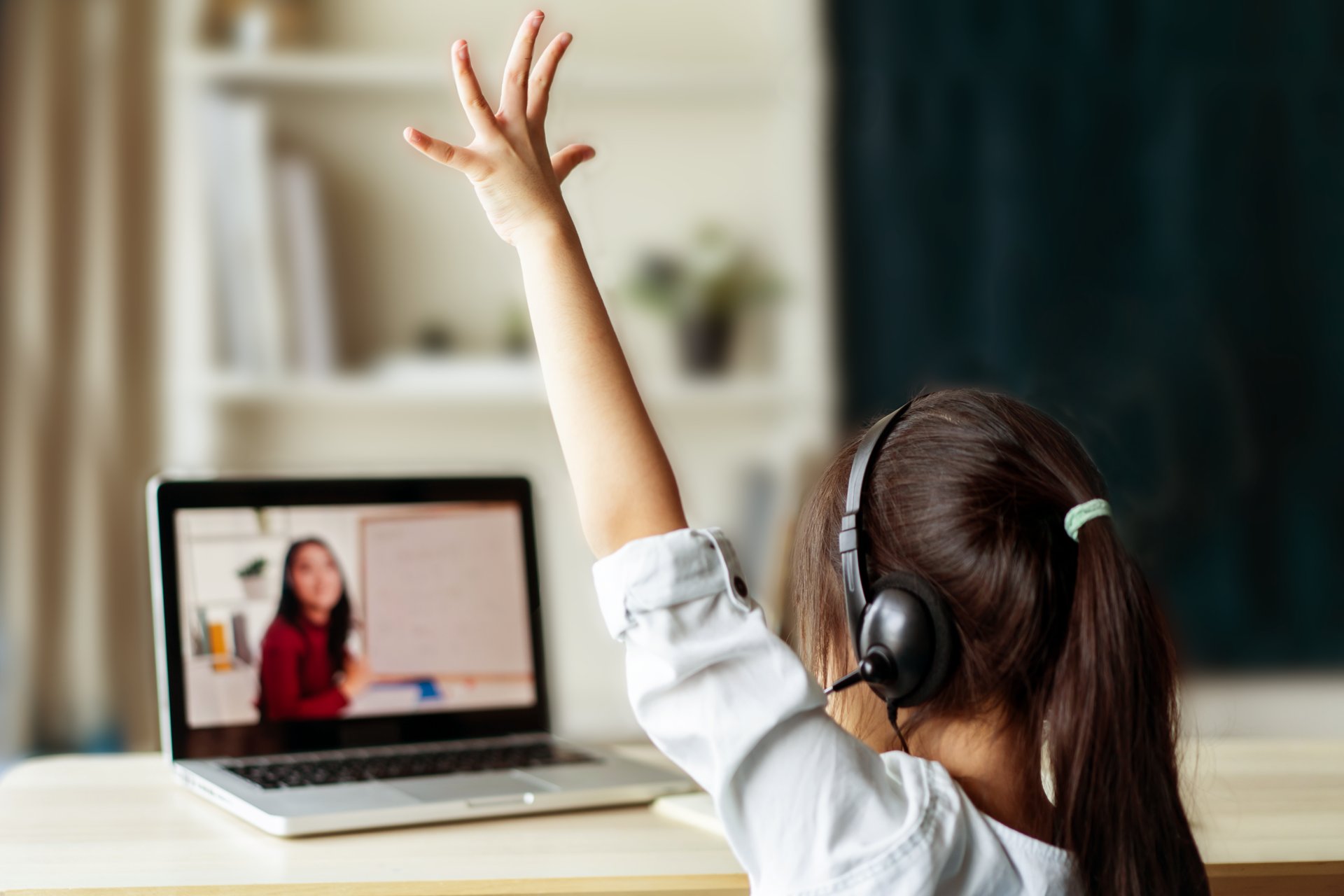 Young student wearing headphones raises hand while attending an online class on a laptop, viewed from behind, utilizing the LAUSD Amplify curriculum.