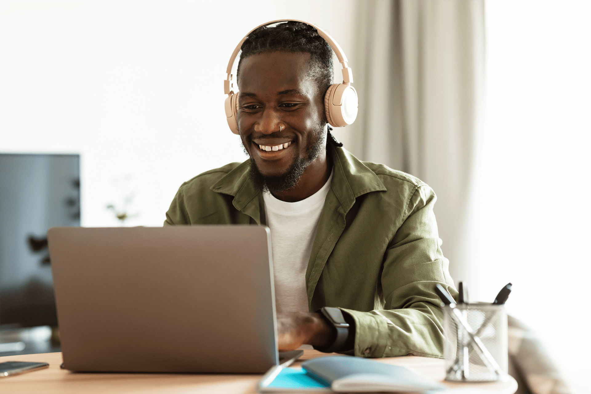 A smiling black man wearing headphones uses a laptop at a desk with office supplies and a monitor in the background, possibly engaging in Amplify Tutoring sessions.