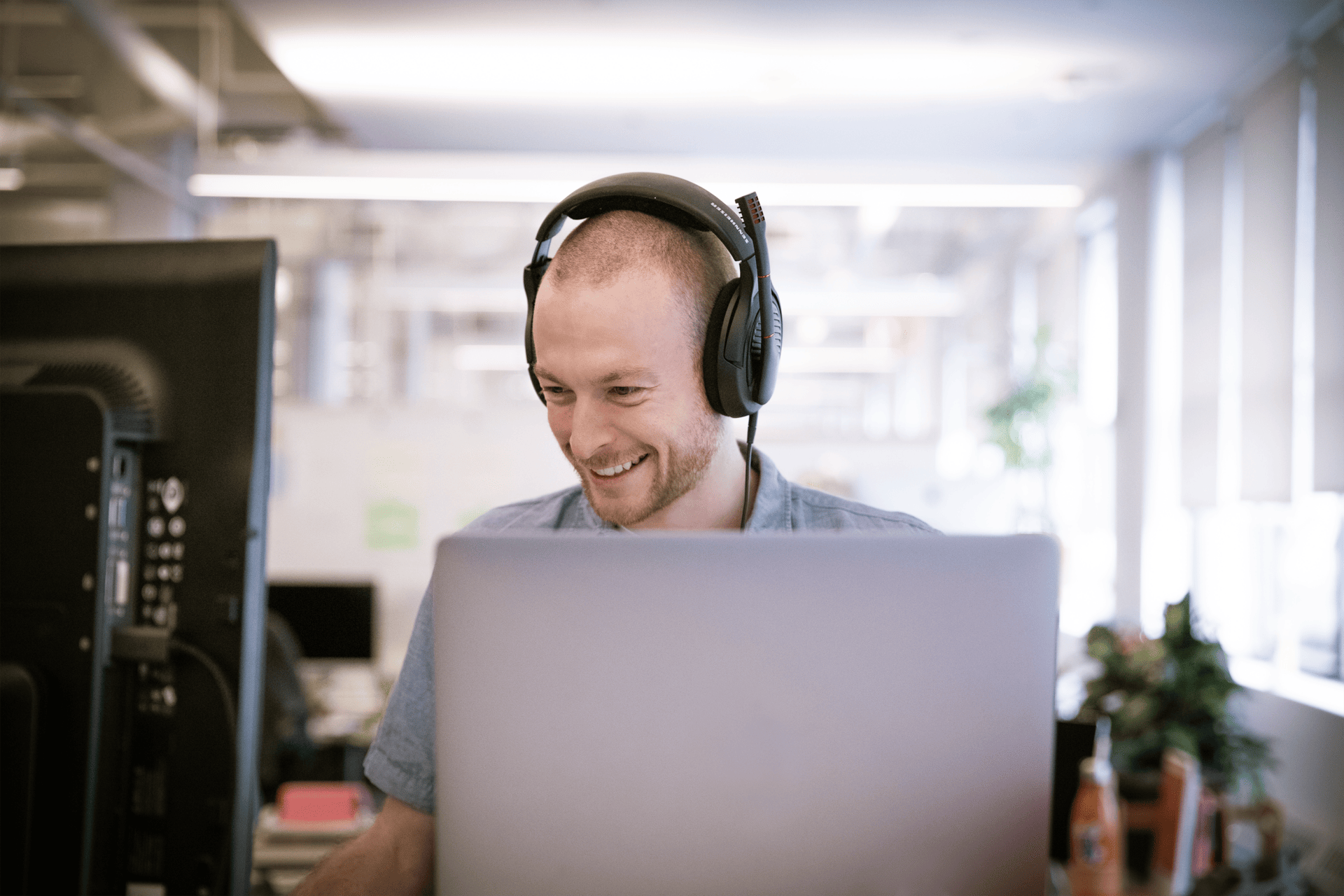 A smiling man wearing headphones and using a laptop in a brightly lit office space for Amplify Tutoring services.